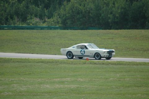 2000 Ford Mustang - A white 1965 Shelby GT350 race car, identified by its distinctive body style and racing decals, navigates a turn on a sunny day.