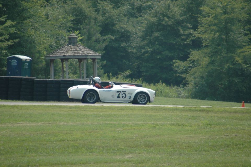 White convertible race car with number 75 on side, driver wearing helmet, on track with trees and gazebo in background
