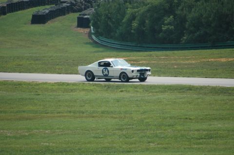 2000 Ford Mustang - A classic white Shelby GT350 race car with blue stripes and the number 54 on its side navigates a turn on a sunny day.