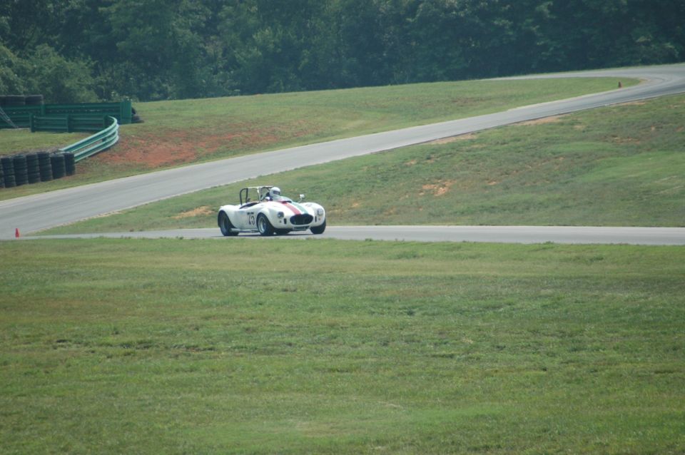 White Cobra-style roadster with red stripe and roll bar racing on a road course