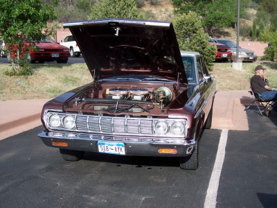 1972 Ford Ranchero - A man sits in a chair next to a classic Plymouth with its hood up, parked in a lot with other vehicles.