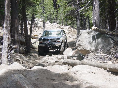 1986 Ford Bronco - A lifted Ford Bronco navigates a challenging rocky path through a forest.