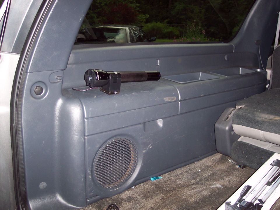 Interior shot of a vehicle's rear storage area, showing a flashlight on a compartment. Another vehicle is visible through the window