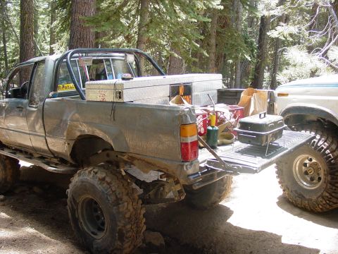 1986 Ford Bronco - Pickup truck loaded with camping gear and tools, parked in a forest setting next to another vehicle.