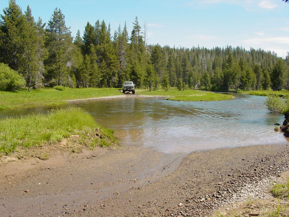 Off-road vehicle crossing a shallow river with trees in background