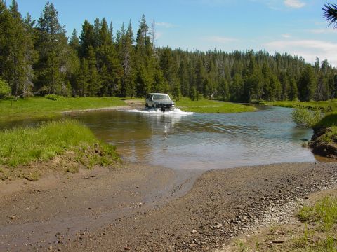 1986 Ford Bronco - A vehicle navigates a water crossing in a wooded area on a sunny day.