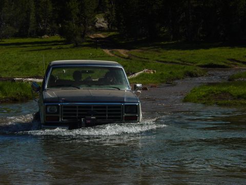 1986 Ford Bronco - A dark blue Ford Bronco navigates a stream on a dirt road, with trees and grassy hills in the background.
