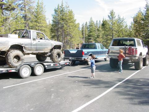 1986 Ford Bronco - Three heavily modified trucks and an SUV are parked in a lot with trees in the background.