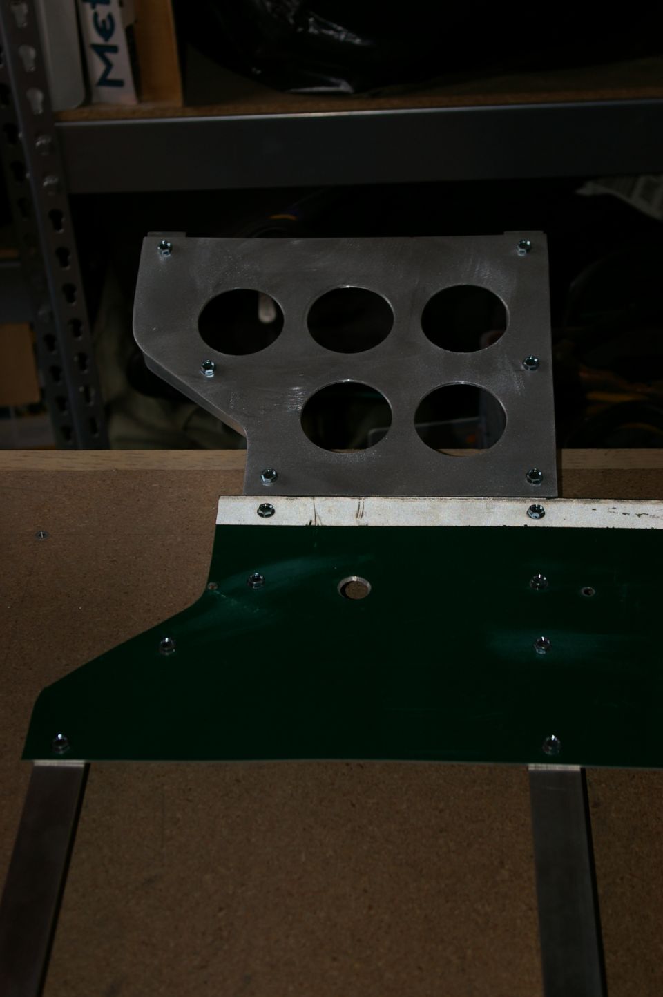 metal bracket with six circular holes, attached to a green panel with screws