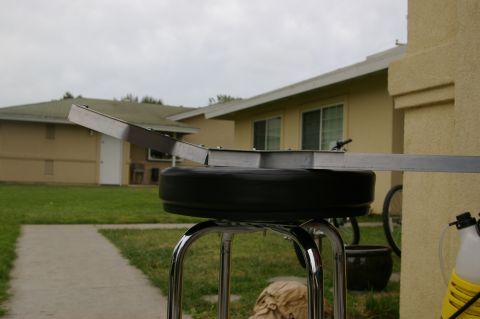 1986 Ford Bronco - A metal bracket is mounted on a chrome stool with a black cushion, set against a backdrop of houses and a cloudy sky.