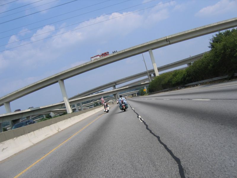 Two motorcycles ride on a highway overpass with multiple concrete ramps overhead