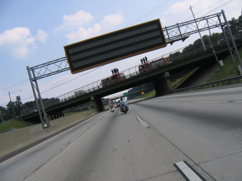 Two motorcycles ride on a highway under an overpass with a large digital sign
