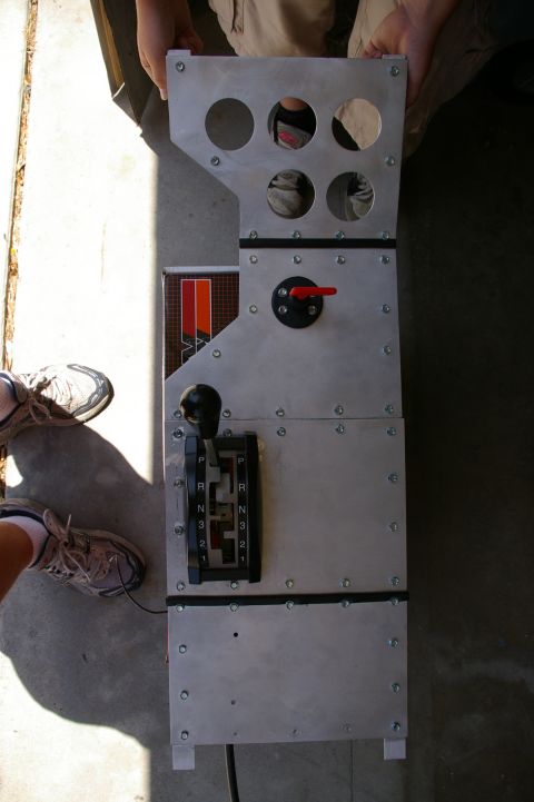 1986 Ford Bronco - An automotive shifter console with multiple circular cutouts and a gear selector is held by two people.