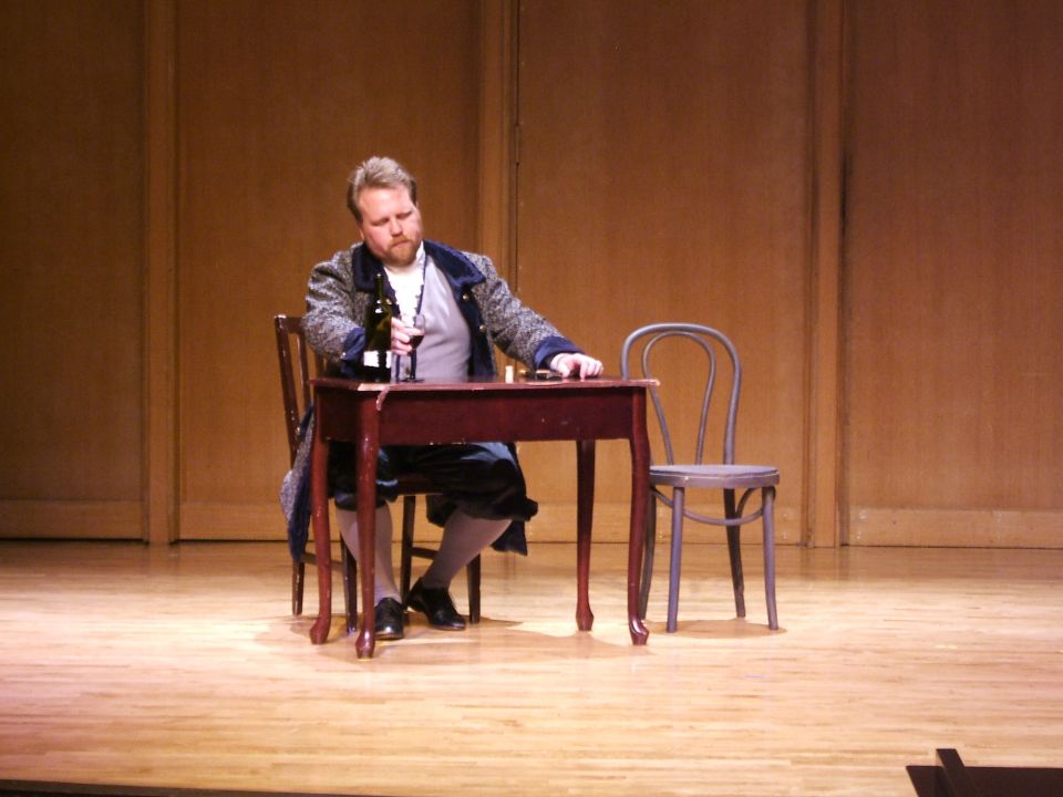 Man in historical costume sits at a wooden table with a wine bottle and glass
