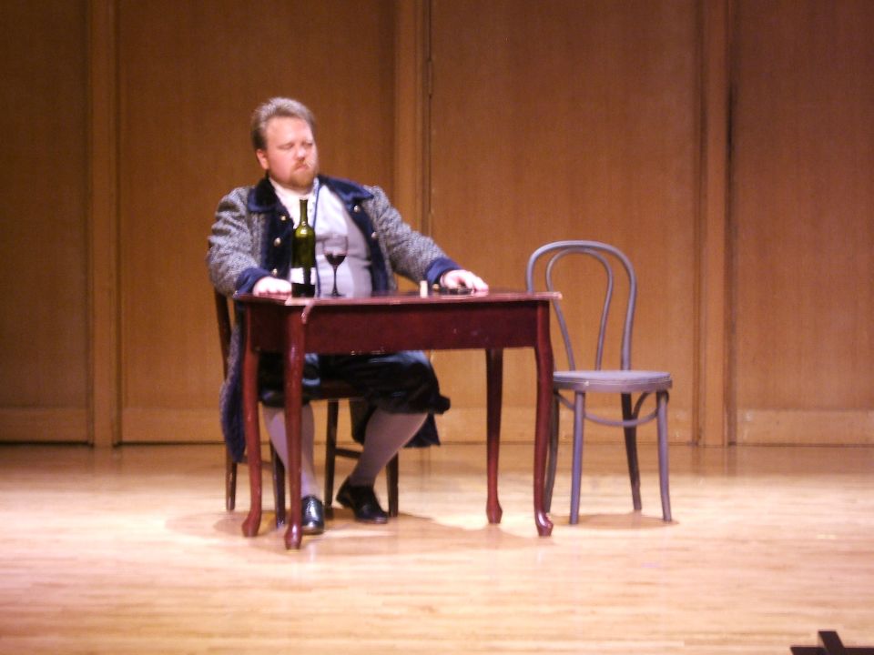 Man in historical costume sits at a table with wine and glass