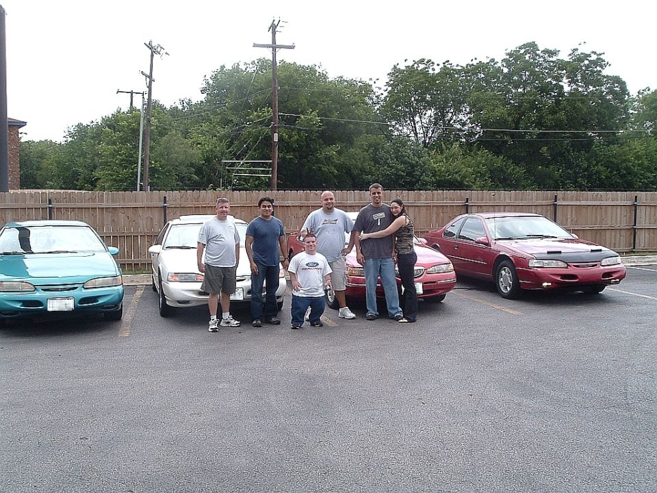 Four cars parked in a lot with a group of people standing in front of them. A teal sedan is on the left, a white sedan is next, a red coupe is in the middle, and a red coupe with hood scoops is on the right