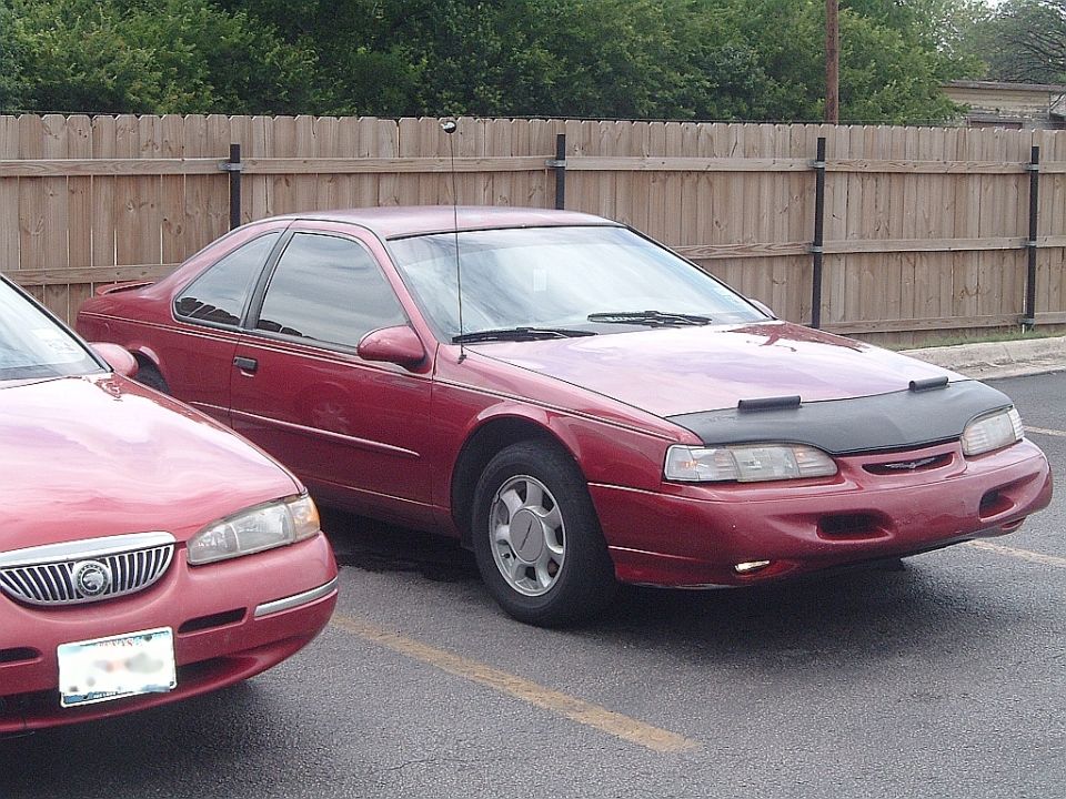 Red coupe with a black car bra parked next to another red car. The coupe has a spoiler and chrome trim