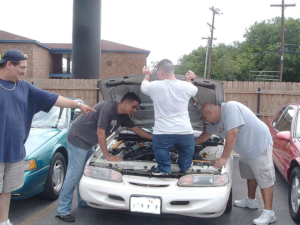 Four men gathered around the open hood of a white car, looking at the engine. Another teal car is visible to the left, and a red car to the right