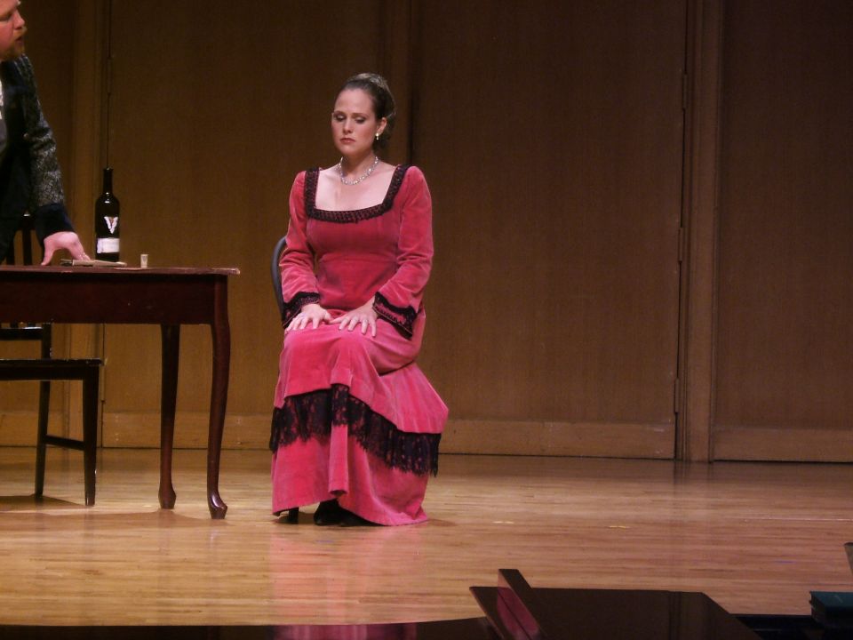 Woman in pink velvet dress with black lace trim sits on stage