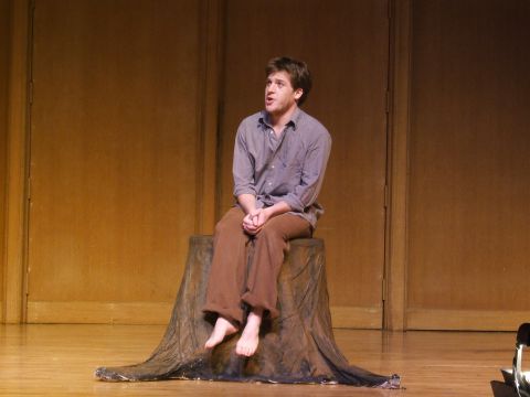 1996 Ford Probe - A man sits barefoot on a stage prop resembling a tree stump, dressed in casual clothing.