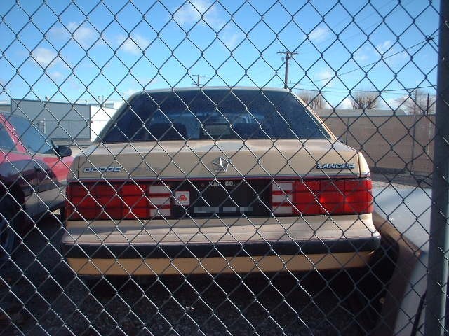Rear of tan Dodge Lancer sedan seen through chain-link fence in a lot