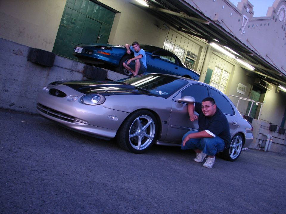 Silver 1998 Mercury Sable with black hood and custom wheels, front 3/4 view, with a man crouching beside it
