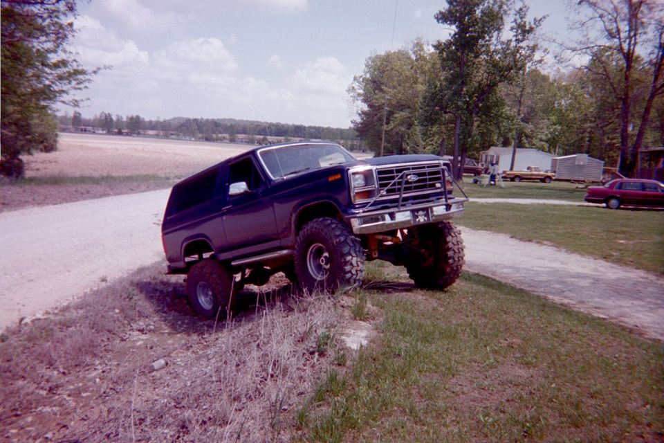Lifted dark blue Ford Bronco with big off-road tires parked on a grassy mound beside a dirt road