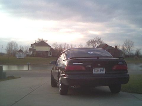 1993 Ford Taurus - The rear of a black sedan is visible with a license plate reading 3284199 under a cloudy sky.