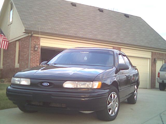 Dark Ford sedan parked in driveway, 3/4 front view, brick house behind