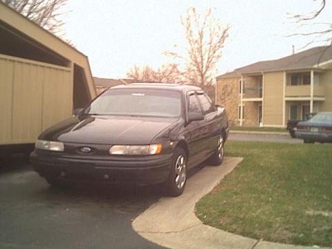 1993 Ford Taurus - A black sedan is parked on a concrete driveway next to a building.