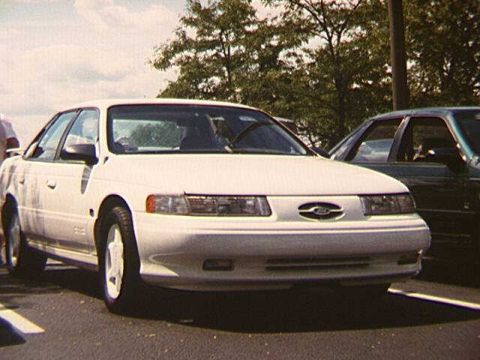 1993 Ford Taurus - A white sedan is parked on asphalt, with trees and a cloudy sky in the background. Another vehicle is visible to the right.
