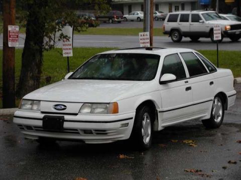 1993 Ford Taurus - A white sedan, likely a 1992 model, is parked on a wet road with other vehicles in the background.