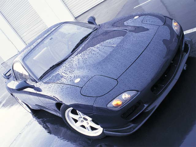 Water-beaded dark blue Mazda RX-7 FD with pop-up headlights up and white wheels, high angle