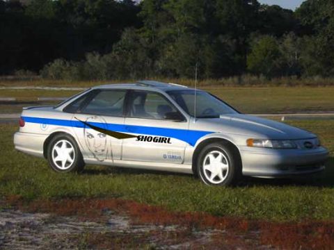 1993 Ford Taurus - A silver 1993 Ford Taurus sedan is shown from a three-quarter front view, custom blue racing stripes and a graphic of a woman's face.