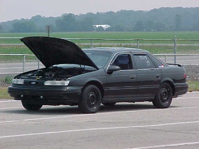 1993 Ford Taurus - A dark sedan sits on asphalt with its hood open, showing the engine bay.