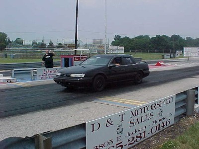 1993 Ford Taurus - A black sedan sits on a drag strip, with a person visible in the background near a booth.