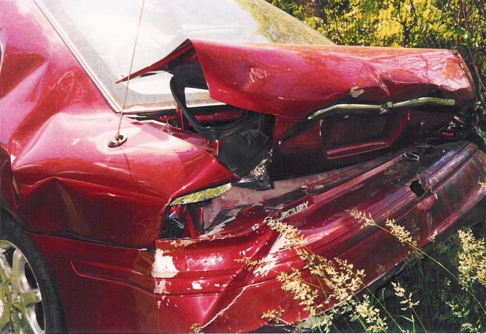 Severely crashed dark red Mercury sedan with crushed rear end in tall grass
