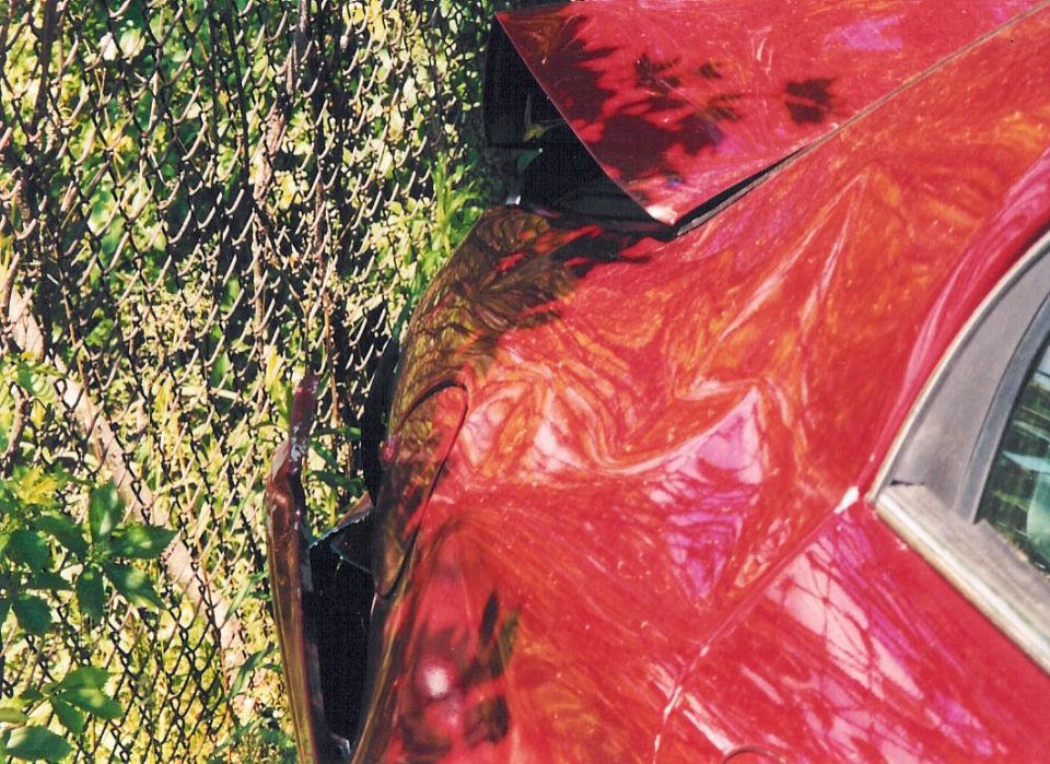 Red sedan rear quarter panel with heavy collision damage against a chain-link fence