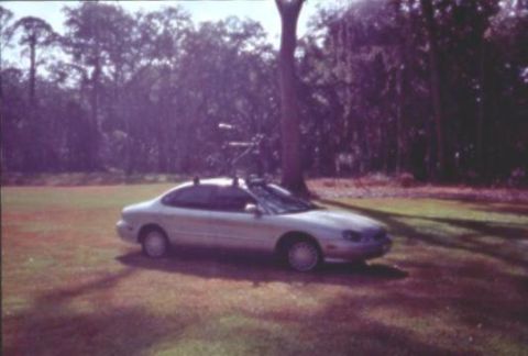 1998 Ford Taurus - faded photo of a silver four-door sedan carrying a bicycle on a roof rack, parked on grass beside wooded ground.