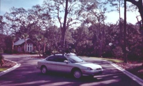 1998 Ford Taurus - silver four-door sedan sits in the parking lot of a small church surrounded by large oak trees, photographed on film.