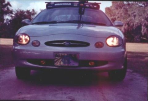 1998 Ford Taurus - Head-on view of a silver third-generation Ford Taurus with headlights and fog lamps lit and a Yakima roof rack, parked on gravel.
