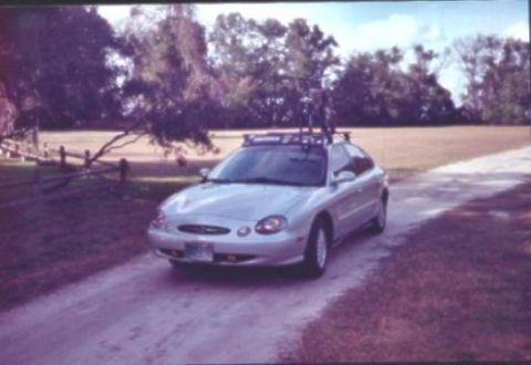 1998 Ford Taurus - A silver Ford Taurus four-door sedan parked at an angle on a dirt road, viewed from the front and passenger side, with a roof rack carrying…