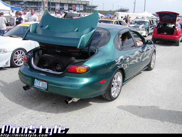 Teal Taurus at car show, trunk open showing subwoofer install and ghost flames