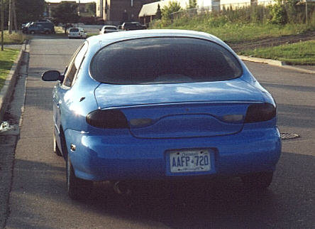 1998 Ford Taurus - Rear view of a bright blue late-1990s Ford Taurus sedan parked at the curb on a residential street, showing the wraparound one-piece tail l…