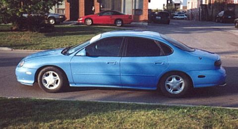1998 Ford Taurus - A blue late-1990s Ford Taurus sedan photographed in side profile at a residential curb. It has tinted windows, body-color mirrors, and mach…
