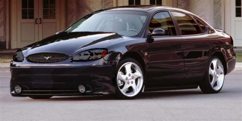 1998 Ford Taurus - Dark plum/near-black Ford Taurus sedan photographed from a low front three-quarter angle in front of a light brick building. It wears a mes…