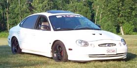 1998 Ford Taurus - Low-resolution three-quarter front view of a white late-1990s Ford Taurus sedan with a windshield banner reading STORM, a raised hood scoop…