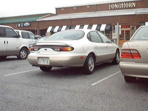 1998 Ford Taurus - A two-tone Ford Taurus sedan, white roof and upper body over gold lower panels, parked rear-first outside a Longhorn restaurant, seen from…