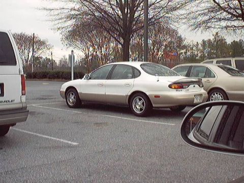 1998 Ford Taurus - A silver-champagne late-1990s Ford Taurus sedan shown from the rear three-quarter in a parking lot, fitted with a raised trunk spoiler and…
