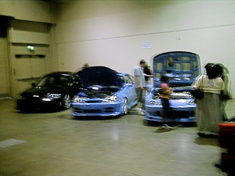 1998 Ford Taurus - Several custom cars are displayed indoors with their hoods open, attracting attention from onlookers.